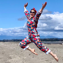Load image into Gallery viewer, Woman wearing colourful floral overalls jumping on a beach in New Zealand with blue sky and mountains in the background.