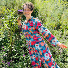 Load image into Gallery viewer, Woman in a colourful floral jumpsuit standing in a garden holding an artichoke flower.
